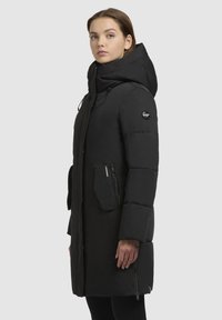Schwarze Daunenjacke mit hohem Kragen und angebauter Kapuze. Mit Reißverschlusstaschen und einem leicht oversized Schnitt. Glatte, matte Textur.