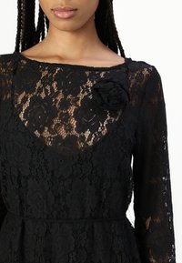 Μαύρο δαντελένιο φόρεμα με μακριά μανίκια, λουλουδάτο μοτίβο, στρογγυλή λαιμόκοψη και μια μαύρη υφασμάτινη ανθοδέσμη στον αριστερό ώμο.