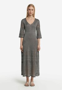 Grå stickad maxiklänning med V-ringning, trekvartsärmar och ett texturerat mönster. Kombinerad med vita remsandaler.