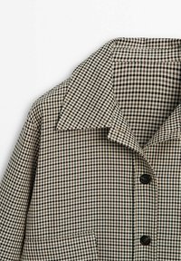 Manteau à carreaux noir et blanc avec un large col et deux boutons noirs visibles à l'avant sur fond blanc.