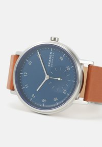 Skagen-klocka med en rund blå urtavla, silverfärgad metallkapsel och brunt läderarmband. Har minimalistiska tim- och minutvisare samt subtila markeringar.