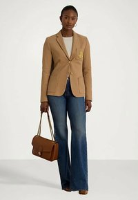 Žena u bež sakou, bijeloj majici, plavim širom farmerkama, smešnim potpeticama, koja nosi smešnu torbu s zlatnim lančićem.