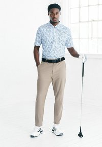 Jasnoniebieska koszulka polo w wzory, beżowe spodnie, czarny pasek, białe buty golfowe z czarnymi paskami, trzymający kij golfowy, stojący w luźnej pozie.