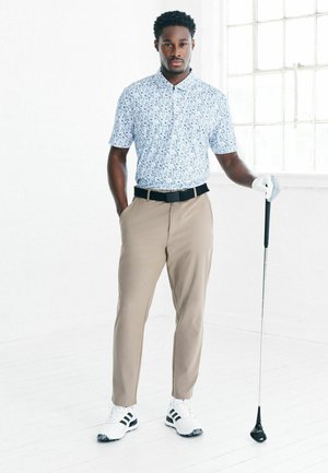 Heled sinine mustriga polo, khaki püksid, must vöö, valged golfijalatsid mustade triipudega, hoiab golfikeppi, seisab lõdvestunult.