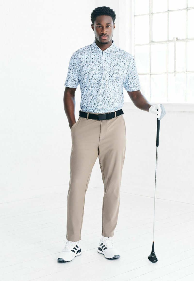 Hellblau gemustertes Poloshirt, khakifarbene Hose, schwarzer Gürtel, weiße Golfschuhe mit schwarzen Streifen, hält einen Golfschläger, steht lässig.