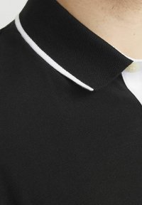 Zwarte polo met een textuur stof, een klassieke kraag met witte rand en een enkele knoopsluiting bij de hals.