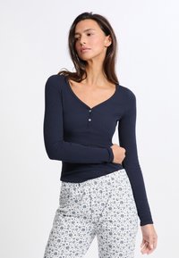 Námořnická dlouhá halenka s výstřihem do V a třemi knoflíky, kombinovaná s bílými vzorovanými kalhotami s modrými akcenty a vzory.