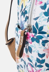Sac à motifs floraux avec un fond blanc, une fermeture éclair métallique, des accents marron et une texture avec des feuilles et des fleurs colorées.