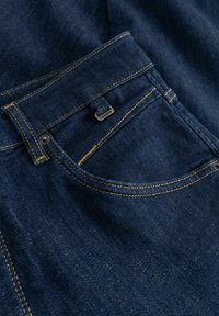 Buzunar de blugi din denim albastru închis cu cusături galbene și o mică etichetă metalică inscripționată cu "GABBA" pe buzunarul pentru monede.