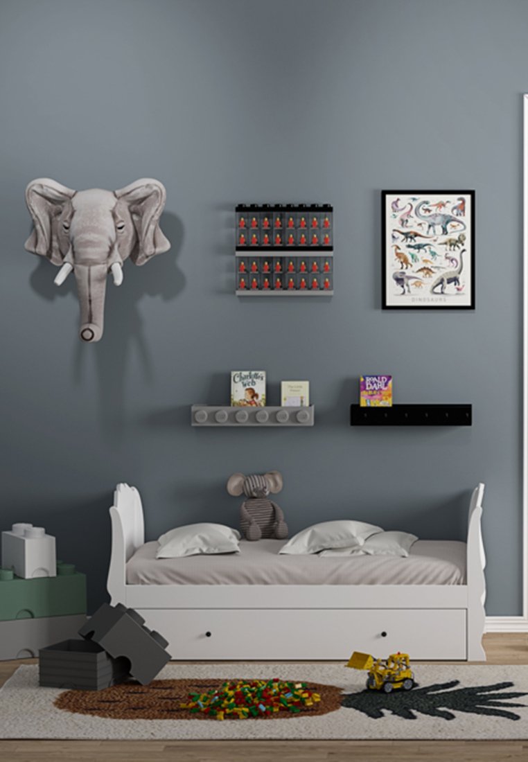 Weißes Holzbett mit grauer Bettwäsche, plüschigem Elefanten-Dekor, grauer Wand, Regale mit Spielzeug, Kinderbüchern und einem gemusterten Teppich mit bunten Blöcken.