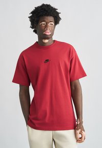 Κόκκινο βαμβακερό μπλουζάκι με στρογγυλή λαιμόκοψη, κοντά μανίκια και μαύρο λογότυπο Nike στο κέντρο του στήθους. Φορεμένο με μπεζ παντελόνια.