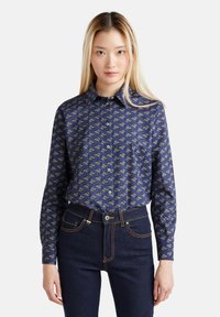 Camicia blu navy con motivo floreale in oro e crema. Maniche lunghe, colletto e una texture liscia. Abbinata a jeans in denim scuri.