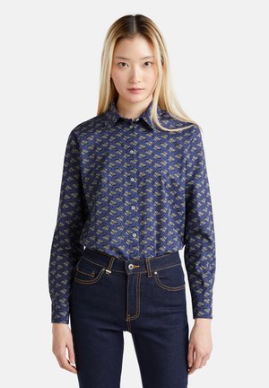 Camicia blu navy con motivo floreale in oro e crema. Maniche lunghe, colletto e una texture liscia. Abbinata a jeans in denim scuri.