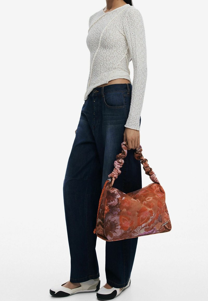 Femme portant un haut blanc texturé à manches longues, un jean sombre ample, des chaussures blanches et noires, tenant un sac à main orange et rose à motif avec anse froncée.