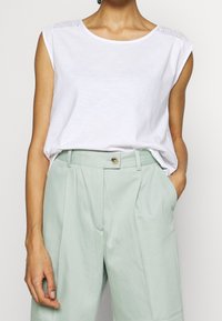 Top blanco sin mangas con un escote redondo y detalles de ojales en los hombros, combinado con pantalones de tiro alto en un tono verde claro con bolsillos.