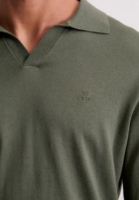 Polo vert olive en mélange de coton avec un col en V et des manches courtes, comportant un logo brodé discret sur la poitrine. Texture lisse.