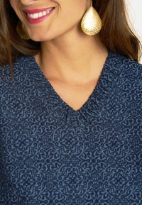 Marineblaues V-Ausschnitt-Strickoberteil mit aufwendigem Muster und strukturiertem Stoff; verfügt über eine geraffte Detail am Hals und wird durch große goldene Ohrringe akzentuiert.