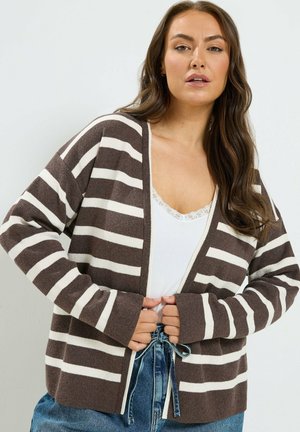 Kvinde iført en brun og hvid stribet cardigan over en hvid top med blonder og blå jeans, som holder i kanterne af cardiganen.