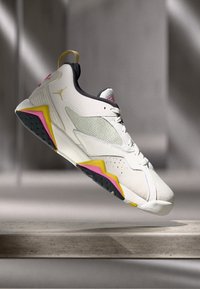 Λευκό αθλητικό sneaker με μαύρες, κίτρινες και ροζ λεπτομέρειες, κρεμασμένο πάνω από μια πέτρινη επιφάνεια σε θολό γκρίζο φόντο.