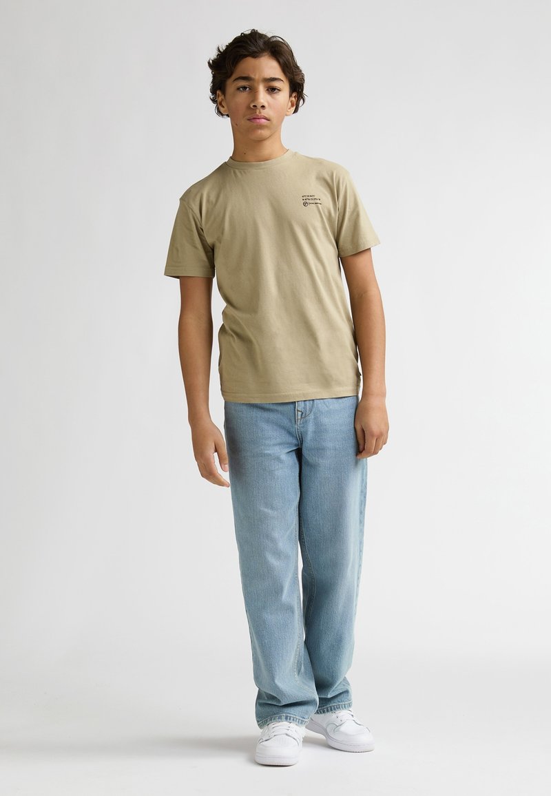 T-shirt en coton kaki à manches courtes, avec un petit motif graphique. Associé à un jean denim bleu clair et des baskets blanches. Fond uni.