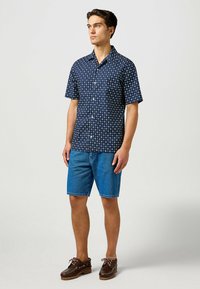 Camisa de manga corta azul marino con patrones circulares blancos, combinada con pantalones cortos de mezclilla azules y zapatos de barco marrón oscuro. Diseño simple y casual.