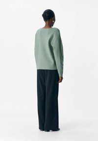 Persoon met een donkere huid en kort haar staat met de rug naar ons toe, gekleed in een losse lichtgroene sweater en brede zwarte broek, tegen een witte achtergrond.