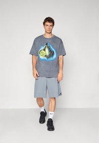 Young man wearing gray wizard graphic T-shirt, light gray knee-length shorts, white Nike socks, and black sneakers, standing against plain background.