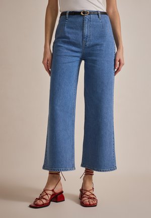 Vaqueros de mezclilla de tiro alto en azul claro con un corte recto y patas anchas. Cuenta con bolsillos delanteros y costuras visibles. Combinados con sandalias de bloque de tacón rojas y con tiras.