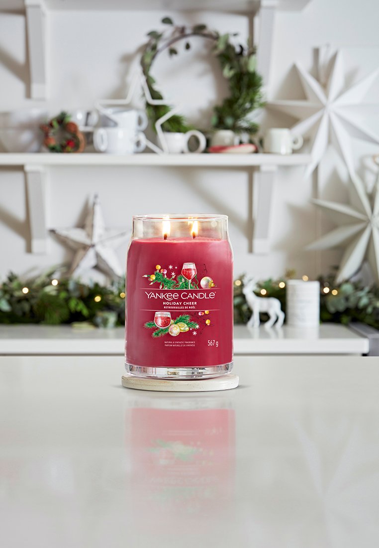 Yankee Candle in einem klaren Glasgefäß, gefüllt mit rotem Wachs, beschriftet mit "Holiday Cheer". Verfügt über zwei Dochte und steht auf einer weißen runden Basis.