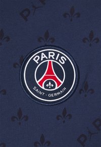 Tela de color navy con un patrón en relieve de flores de lis, que presenta un logo circular del Paris Saint-Germain en el centro con acentos en rojo y blanco.