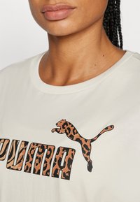 Kreminės spalvos marškinėliai su juodu ir ruduo leopardų rašto logotipu bei ryškiais "PUMA" tekstu leopardų raštu. Lygus audinio paviršius.