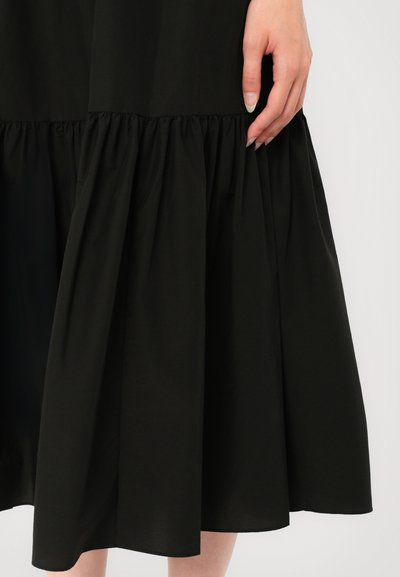 Black, tiered dress with a gathered hem. Made of smooth fabric, it features a fitted bodice and flowing skirt. Hand resting on the fabric.