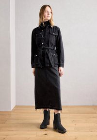 Kobieta ubrana w czarną dżinsową kurtkę z paskiem, czarny golf, długą czarną dżinsową spódnicę z postrzępionym dołem oraz czarne botki na obcasie, stojąca w pomieszczeniu.