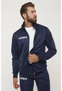 Chaqueta de chándal atlético de color azul marino, hecha de tela suave, con cuello alto, cremallera frontal y logo blanco "LEGEA" en el pecho.