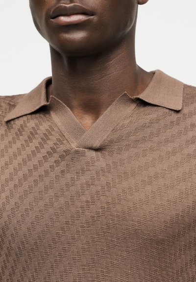 Gros plan sur le bas du visage et le cou d'une personne portant un polo en maille texturée marron clair avec un col en forme de V.