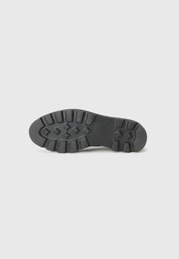 Suela de zapato de goma negra con textura estriada, con tacos y un patrón distintivo de formas elevadas para mayor tracción.