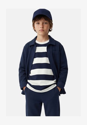 Joven chico con gorra azul marino, chaqueta azul marino y camisa blanca con anchas rayas horizontales azul marino, de pie con las manos en los bolsillos.