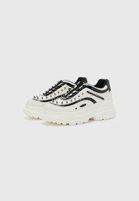 Zapatillas chunky en cuero blanco y negro, con tachuelas metálicas, acentos de cordones texturizados y una suela de goma gruesa. Diseño elegante y llamativo.