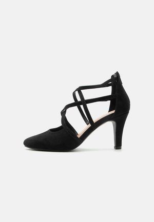 Μαύρο σουέντ ψηλοτάκουνο παπούτσι με μυτερή μύτη και λουριά που διασταυρώνονται στον αστράγαλο, πλευρική όψη σε λευκό φόντο.