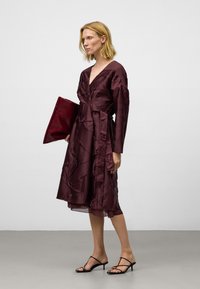 Kvinna i en midilång, djupt burgundy klänning med texturerad tyg, som håller i en stor matchande kuvertväska, iförd svarta klacksandaler med remmar.