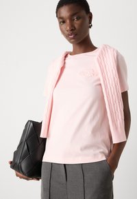 Rosa Baumwoll-T-Shirt mit einem pastellfarbenen Strickpullover, der über den Schultern drapiert ist, und grauen plissierten Hosen. Schwarze strukturierte Handtasche in der linken Hand gehalten.