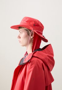 Chapeau de pêche imperméable rouge avec un large bord, une mentonnière ajustable et un logo sur le devant. Assorti avec une veste rouge correspondante.