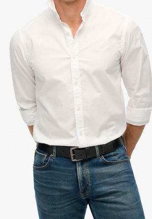 Homme portant une chemise blanche à boutons avec les manches retroussées et un jean bleu maintenu par une ceinture en cuir noir avec une boucle métallique.