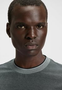 Camicia grigia strutturata con scollatura rotonda, dotata di una fodera interna nera e dettagli di motivo sottili, indossata da un modello con capelli corti.