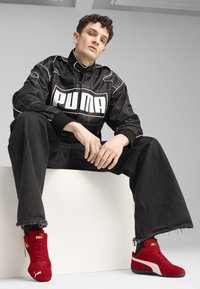Jachetă neagră matlasată cu marele logo alb "PUMA", combinată cu blugi negri largi și adidași roșii din velur, cu accente albe.