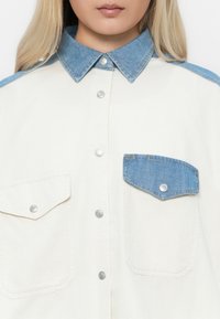 Frau trägt ein weißes Hemd mit blauem Denim-Kragen und Taschenklappen, mit silbernen Druckknöpfen vorne und an den Taschen.
