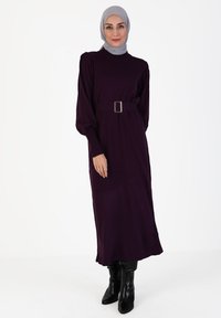Lila Langarmkleid mit hohem Ausschnitt, tailliertem Bund und einem Gürtel mit rechteckiger Schnalle. Getragen mit schwarzen kniehohen Stiefeln.