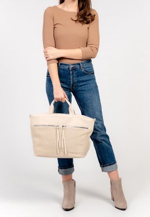 Beige tote bag med struktureret stof og glatte læderdetaljer, der har dobbelte frontlomme med lynlås og håndtaget design, holdt af en person.