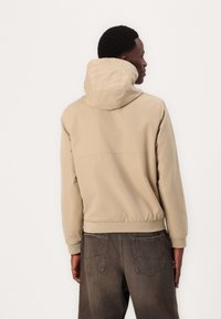 Beige huva jacka med elastisk nederkant och ärmslut. Slät textur, enkel design, har en bakre ok och sitter ovanför mörkgrå jeans.