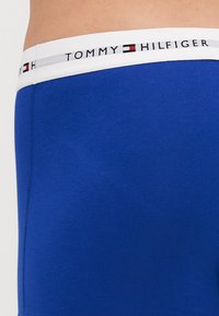 Blå bomulds-boxershorts med en hvid elastisk talje med "TOMMY HILFIGER" logoet og røde og sorte accenter.